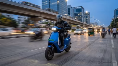 A rider in full gear on a blue Ola S1 Pro Plus electric scooter cruising at dusk on a busy highway with corporate buildings and a metro flyover behind.